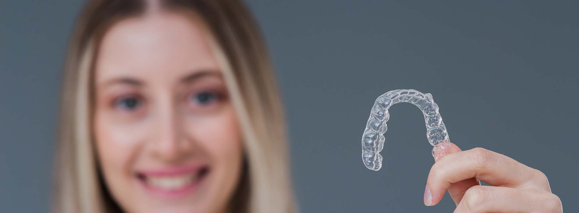 The image features a person holding up a clear plastic object that resembles a mouthguard or dental retainer, with a close-up of the item showing its translucent material and contoured shape, against a blurred background that suggests an indoor setting.