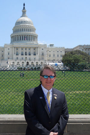 The image features a man standing outdoors in front of the United States Capitol building, wearing a suit and tie with a name tag, posing for the photograph.