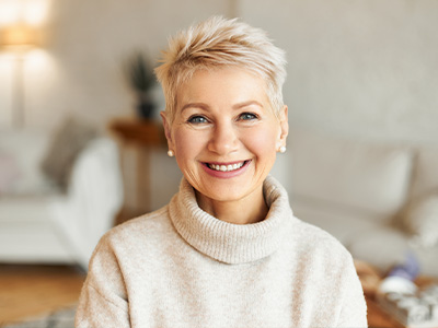 The image features a smiling woman with short hair, wearing a white turtleneck sweater. She appears to be indoors, possibly in a home setting, and there s a warm, inviting ambiance suggesting a comfortable environment.