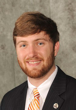 A man wearing a suit and tie, smiling at the camera with a beard, against a wall.