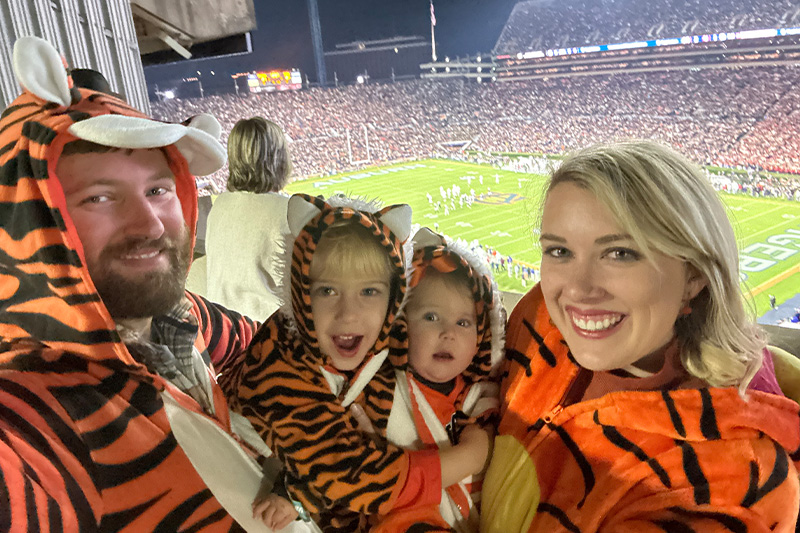 The image shows a family with two adults and two children, dressed in animal costumes, sitting together at a stadium during a football game, wearing matching Tiger outfits and holding up their hands to wave.