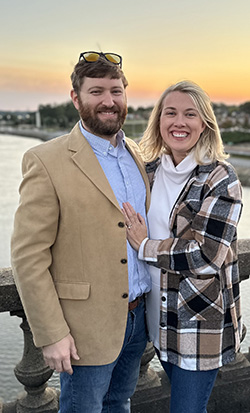 A couple poses together at sunset with a body of water behind them, both smiling and dressed casually.