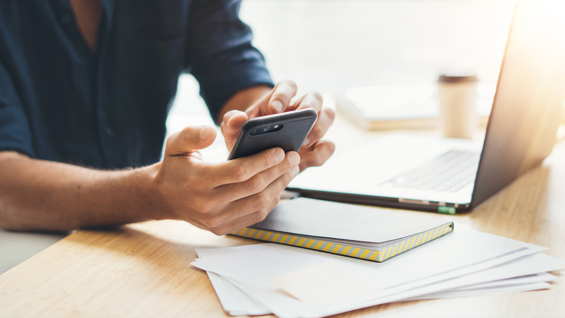 A person s hands are holding a smartphone while sitting at a desk with a laptop and papers.