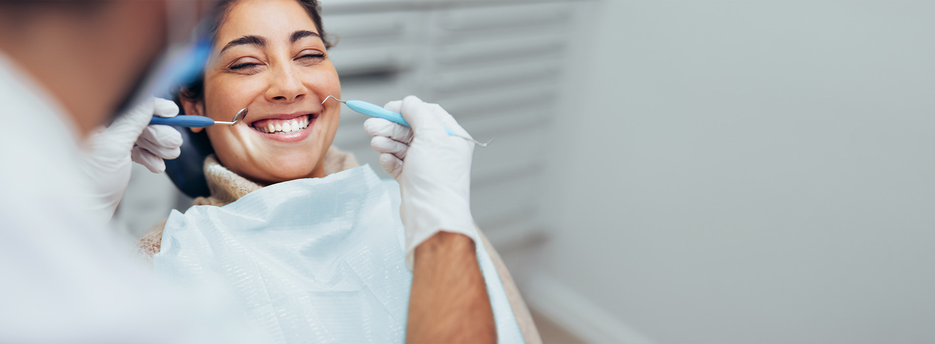 A woman wearing a surgical mask smiles at the camera while sitting in a dental chair with a dentist s hand visible behind her head.