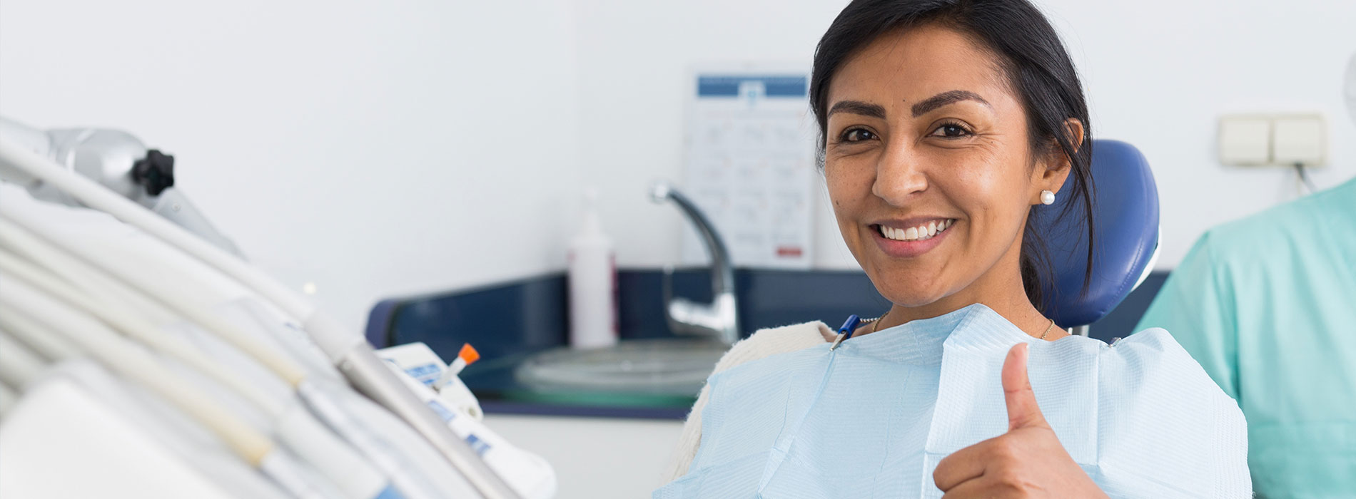 A woman wearing a surgical mask, giving a thumbs-up sign, sits at a dental workstation with medical equipment behind her.