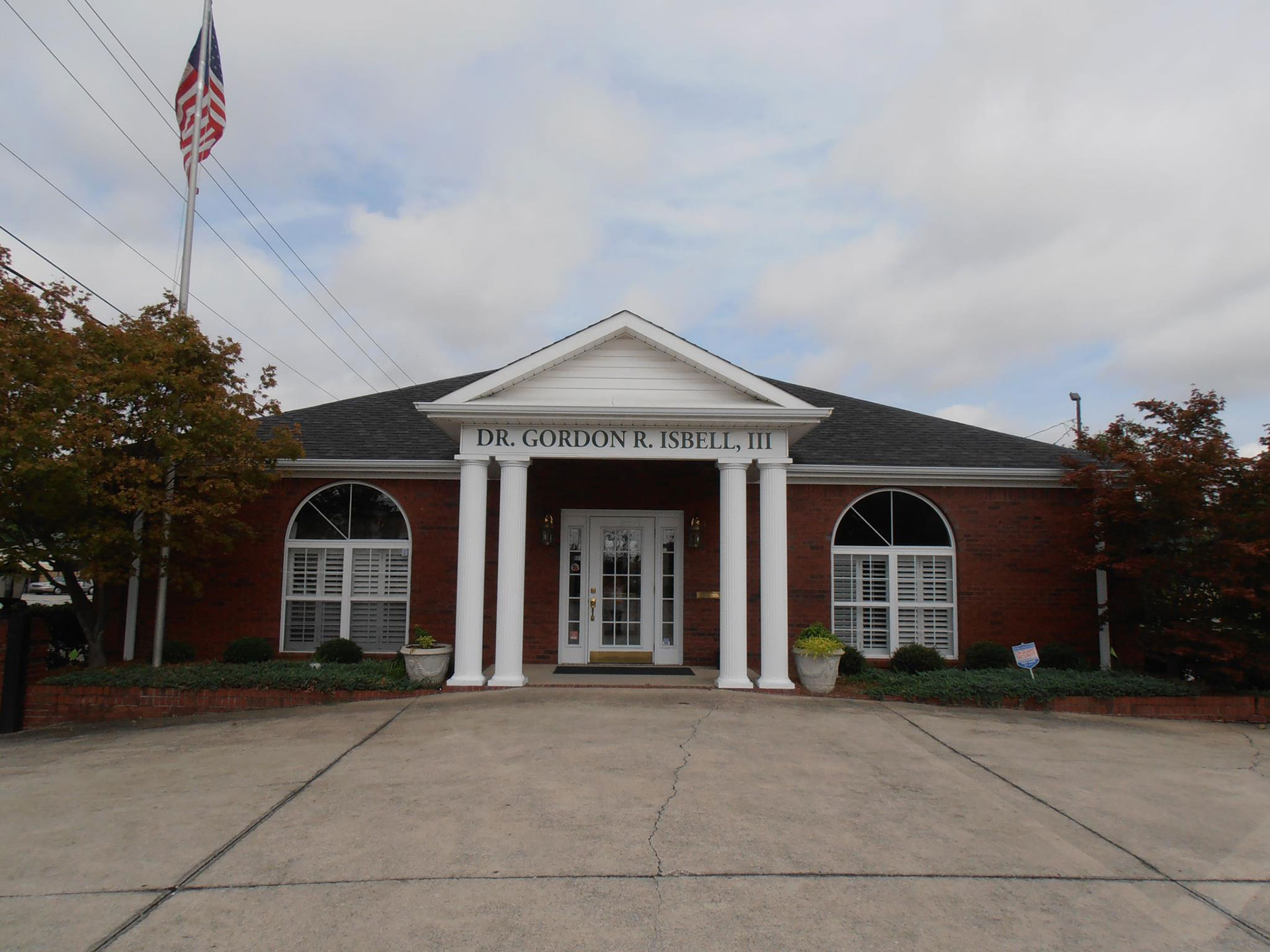 The image shows a building with a sign that reads Dr. Gordon H. Smith III, featuring a red brick facade, white trim, a white roof, a flagpole, and trees in the background.