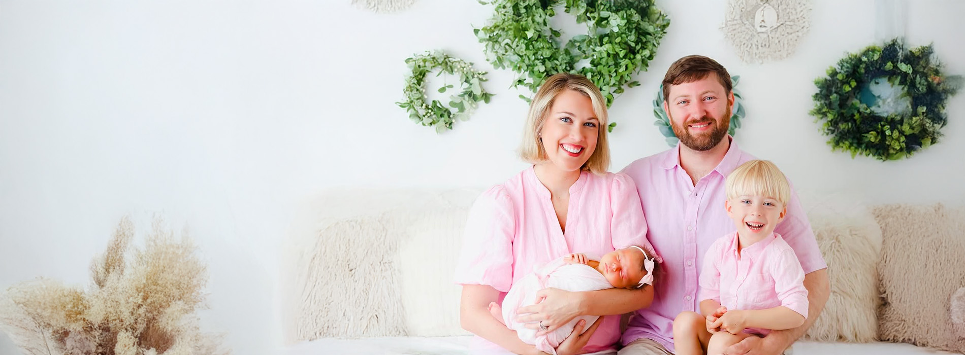A family posing together in front of a white wall with greenery, each member wearing pink shirts.