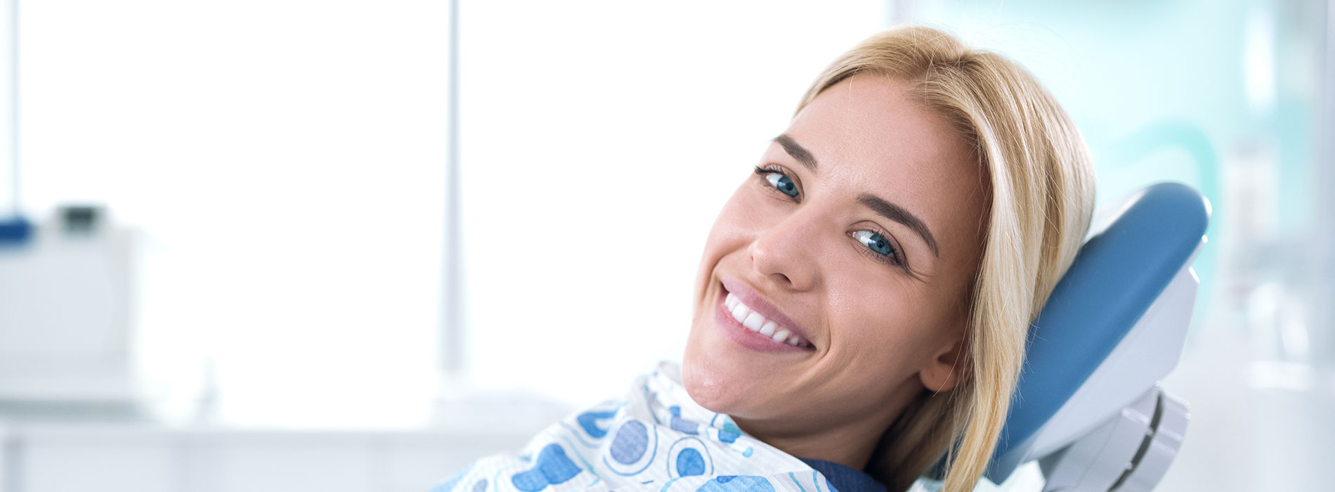 A woman sitting in a dental chair with a smile on her face, wearing a blue shirt, and looking directly at the camera.