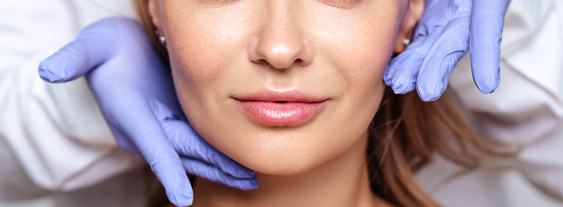 The image features a woman seated with a blue surgical mask covering her face, looking directly at the camera, while another person s hands are gently placed on her cheeks.