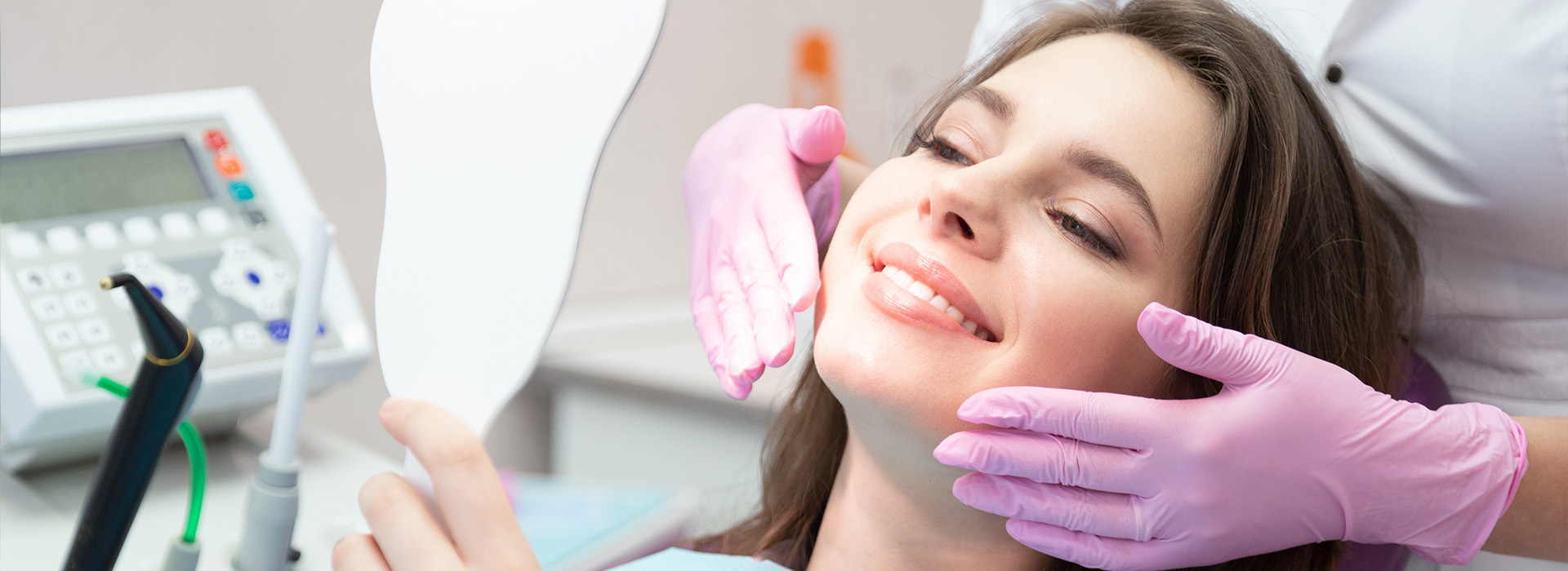 The image shows a woman sitting in a dental chair receiving treatment with a dental hygienist standing behind her, wearing protective gloves and using a device to clean her teeth.