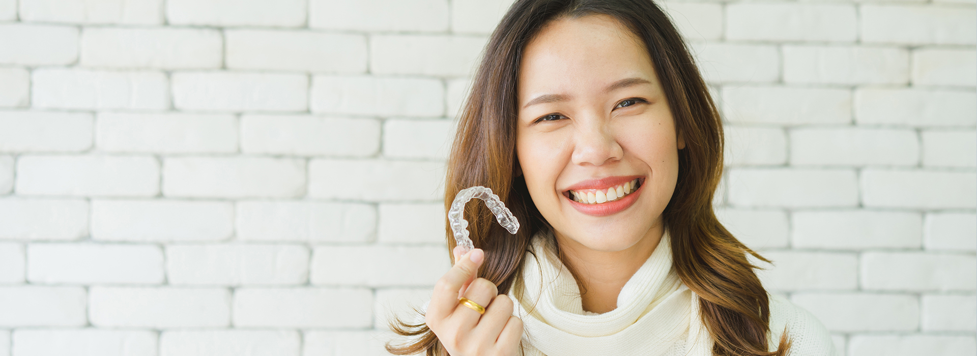 A woman holding a toothbrush with a smile on her face against a brick wall backdrop.