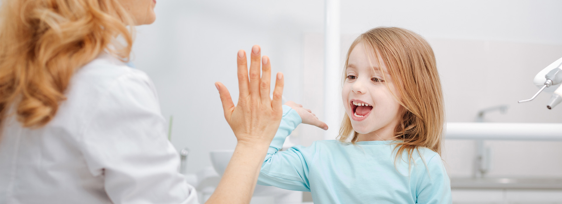 The image shows a young girl waving at someone with her arms raised, standing in front of a woman who appears to be washing her hands. Both are indoors, likely in a bathroom given the presence of a sink and faucet.