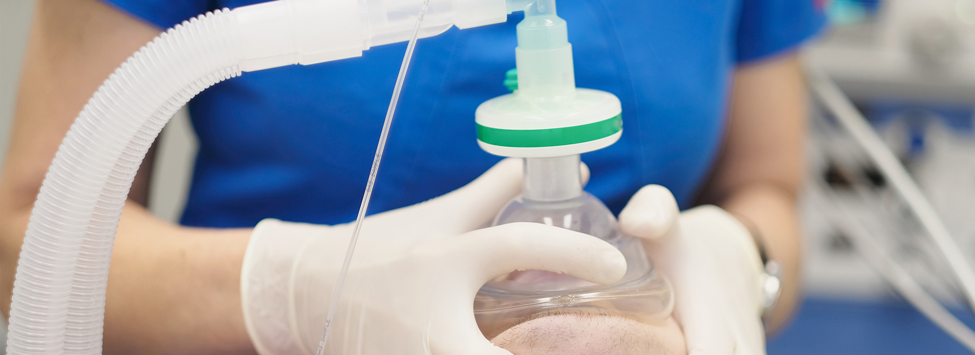 The image shows a medical setting with a person wearing gloves and a surgical mask, holding a clear plastic syringe filled with liquid, which appears to be a medication or solution, while standing near a piece of equipment that could be an IV drip.