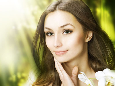A woman with long hair and blue eyes gazes directly at the camera with a soft smile, set against a blurred background of greenery.