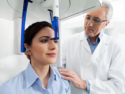 A man standing next to a woman who is seated with her eyes closed under a medical device, possibly for an eye exam or treatment.