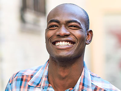 A smiling man with short hair, wearing a blue shirt, stands against a backdrop of a building.