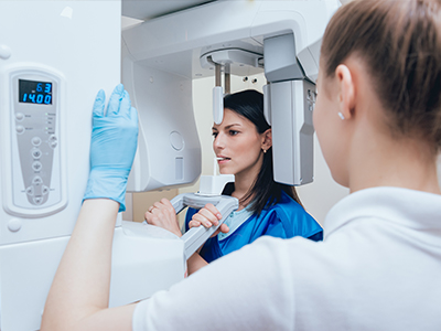 A woman stands next to a large digital mammography machine, both are wearing face masks and gloves, with a blue background featuring a digital display showing 100 and a small screen with an image of a breast.